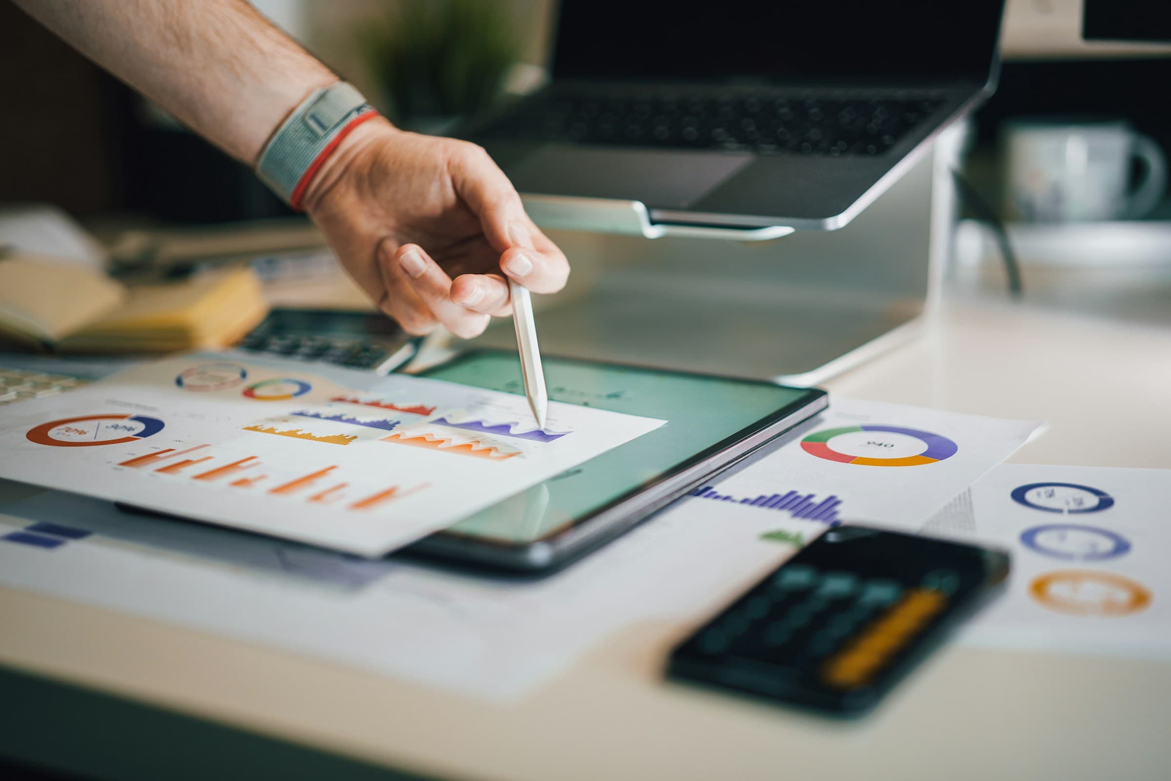 Hand pointing a pen at printed bar charts beside a tablet